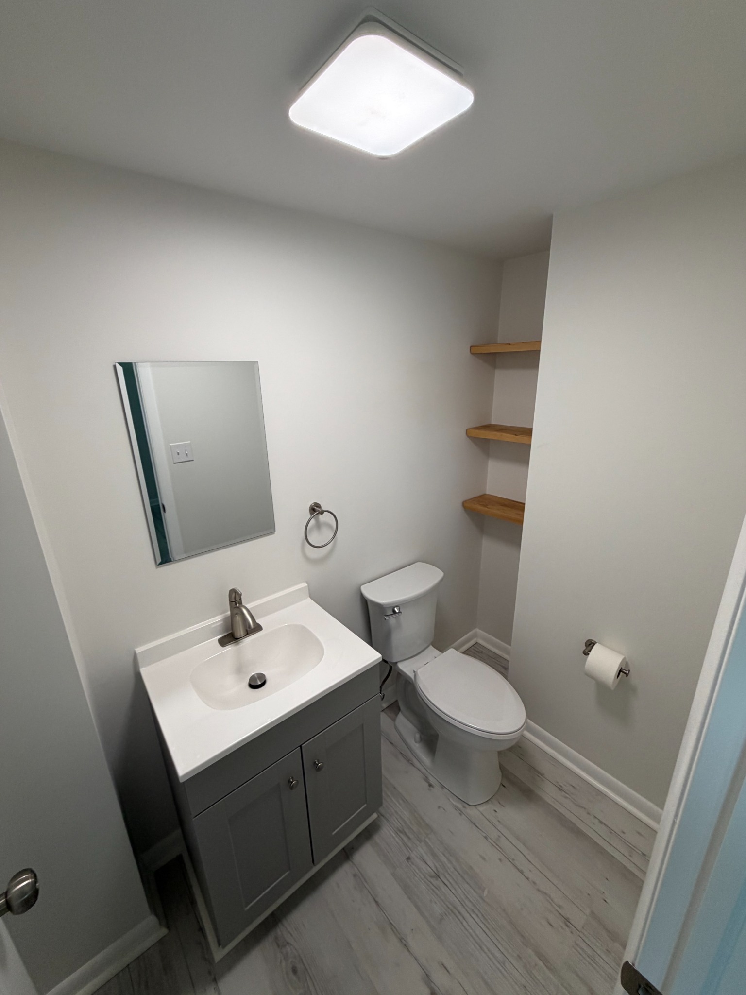 Bright compact powder room with gray vanity, wood shelves and light flooring